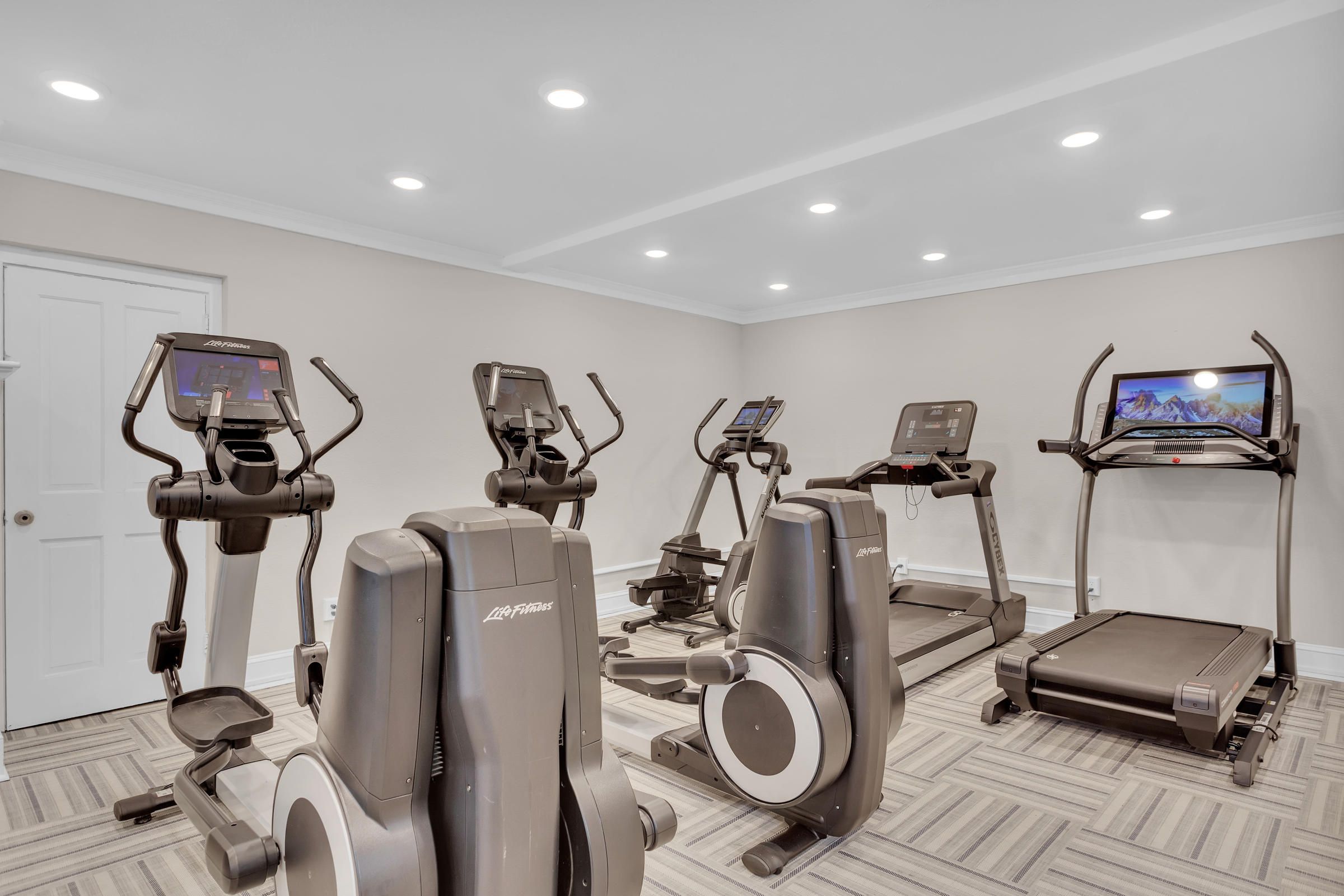 Small gym room with treadmills, ellipticals, and exercise bikes on a patterned floor under bright ceiling lights.