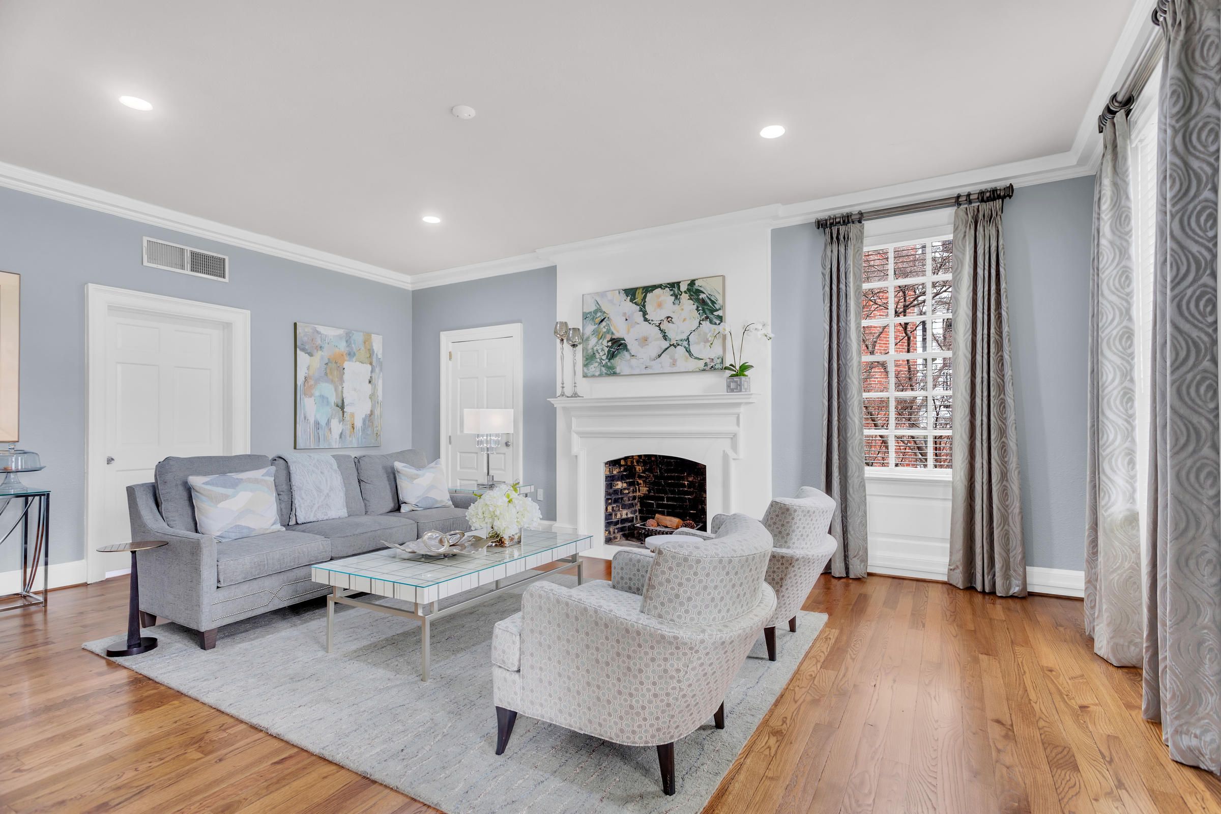 Bright living room with gray furniture, a fireplace, art on the walls, large windows, and hardwood floors.
