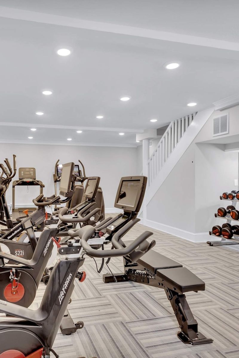 Home gym with exercise bikes, weight bench, dumbbells, and bright lighting next to a staircase.
