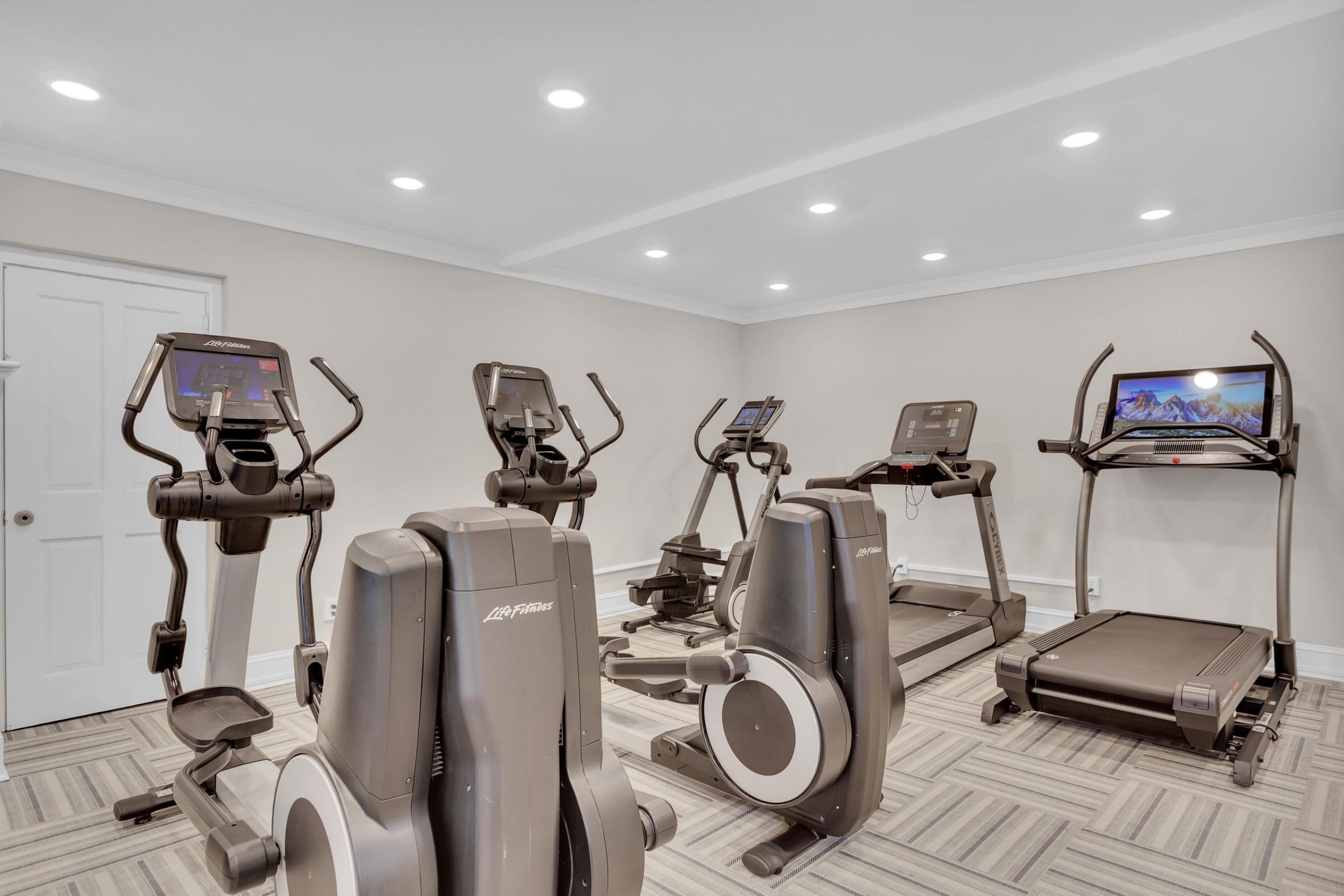 Small gym room with treadmills, ellipticals, and exercise bikes on a patterned floor under bright ceiling lights.