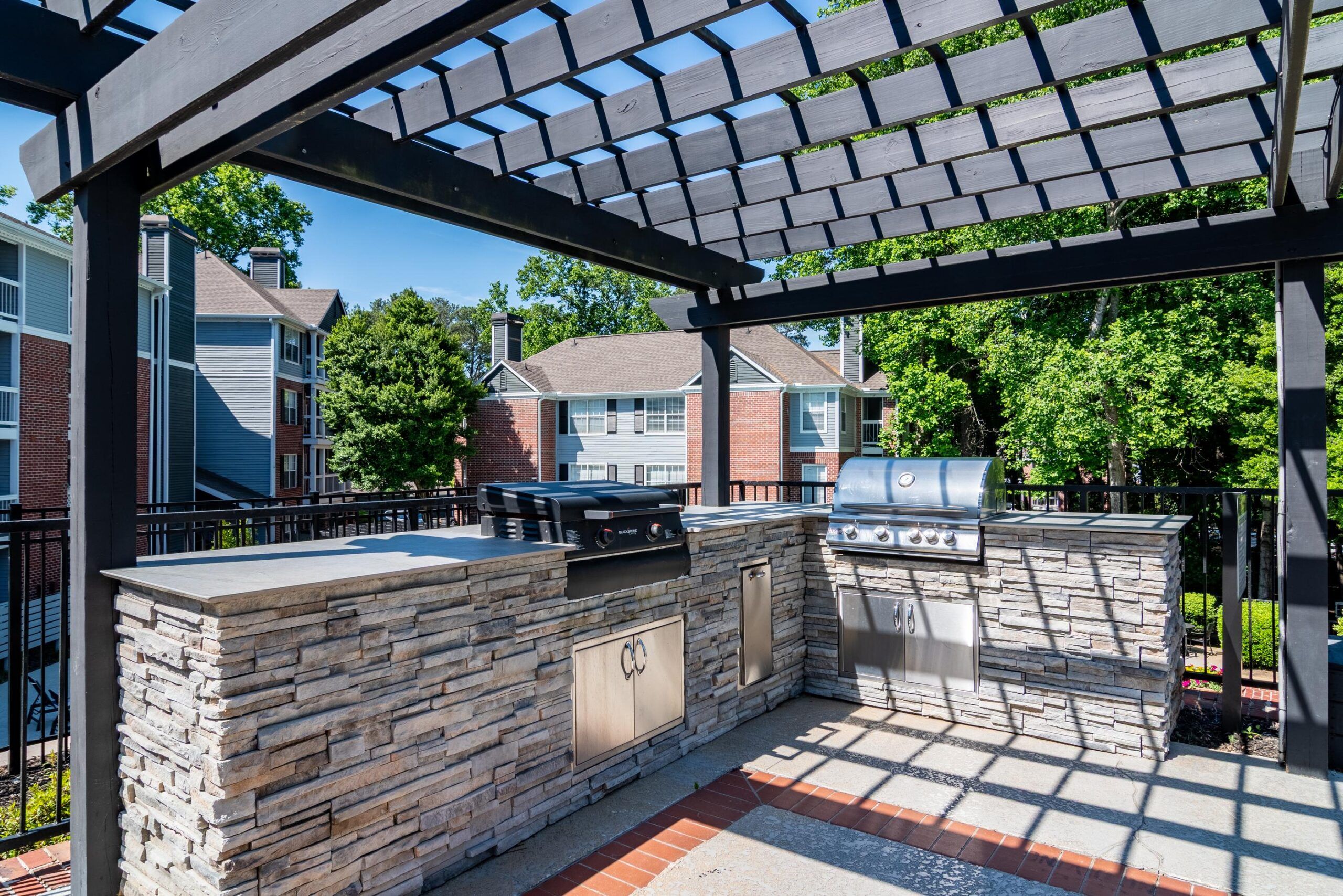 Outdoor kitchen with stone counters, two grills, and a pergola, surrounded by apartment buildings and trees.