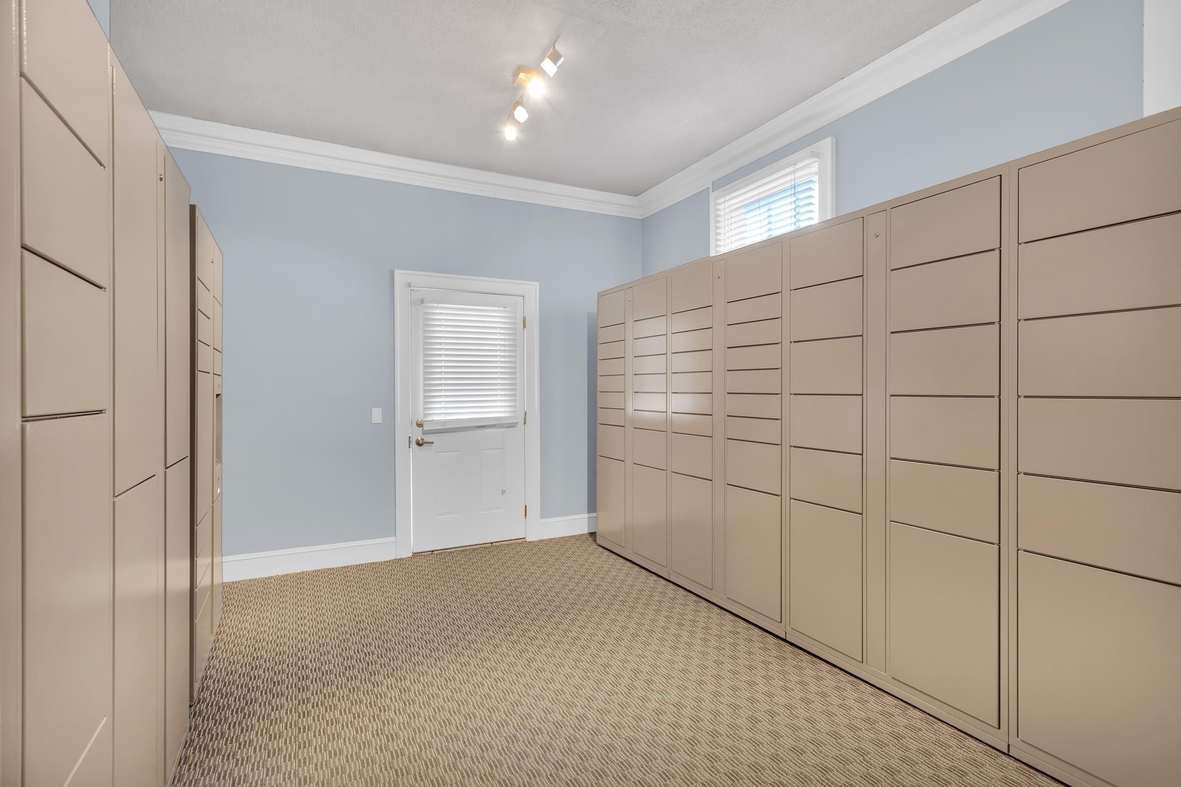 Room with tan metal lockers, beige carpet, blue walls, and a white door and window with blinds.