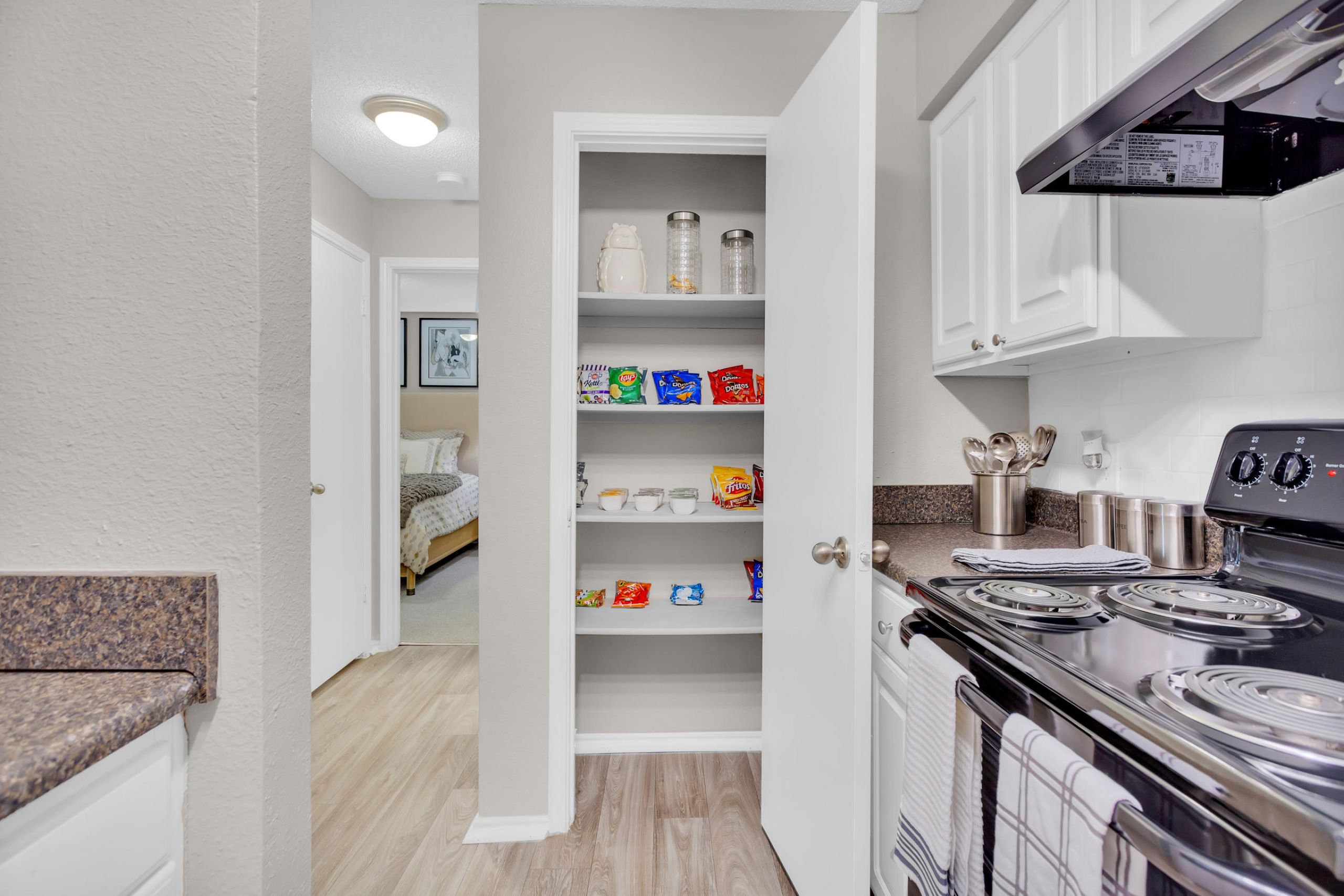 Kitchen with an open pantry stocked with snacks and shelves, next to a stove and hallway to a bedroom.