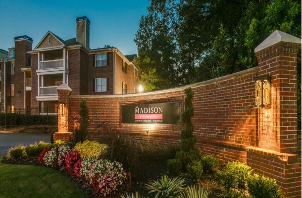 Brick apartment complex at dusk with a lit "Madison Brookhaven Apartment Homes" sign and colorful flowers in front.