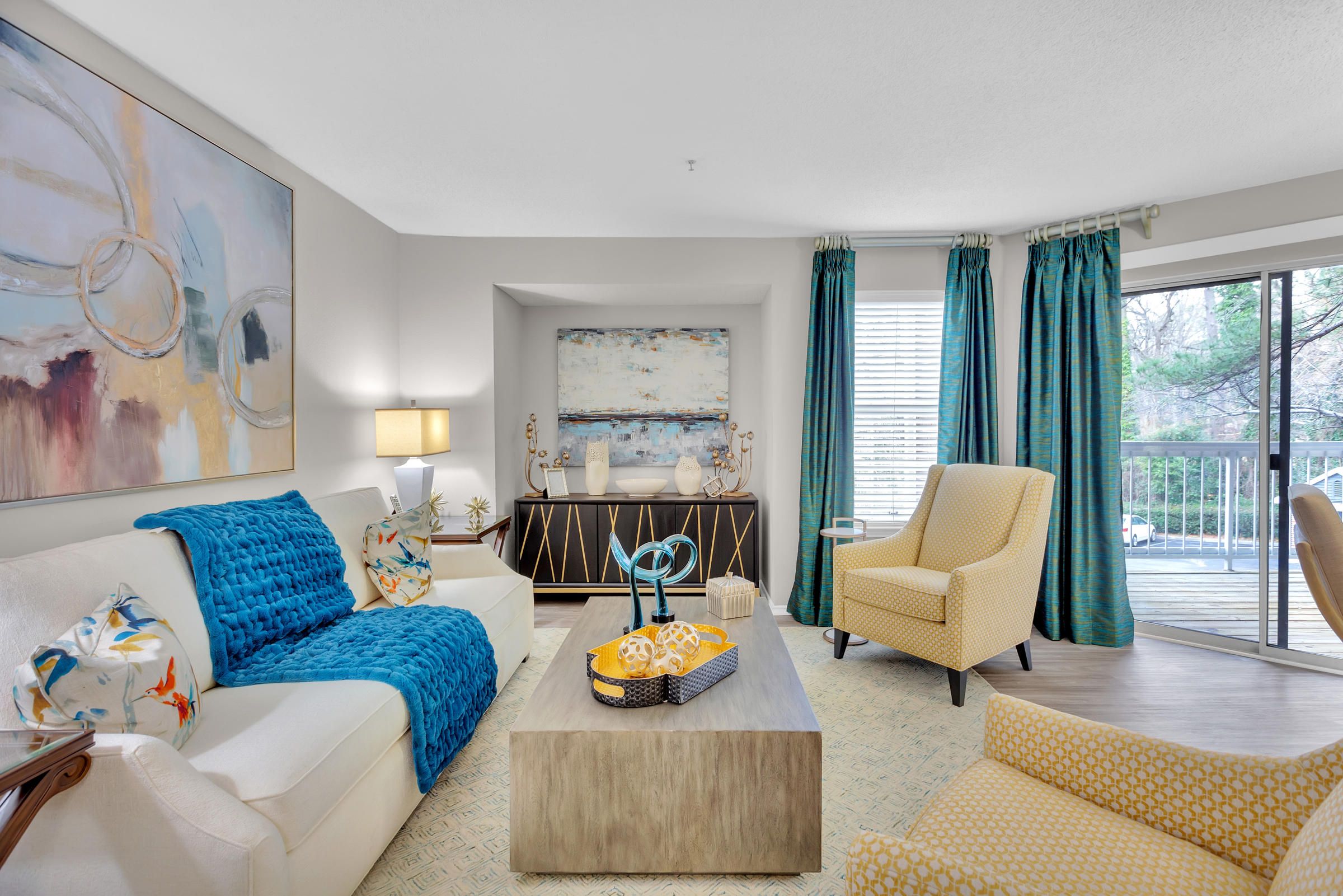 Bright living room with modern furniture, blue accents, abstract art, and large windows facing a balcony.