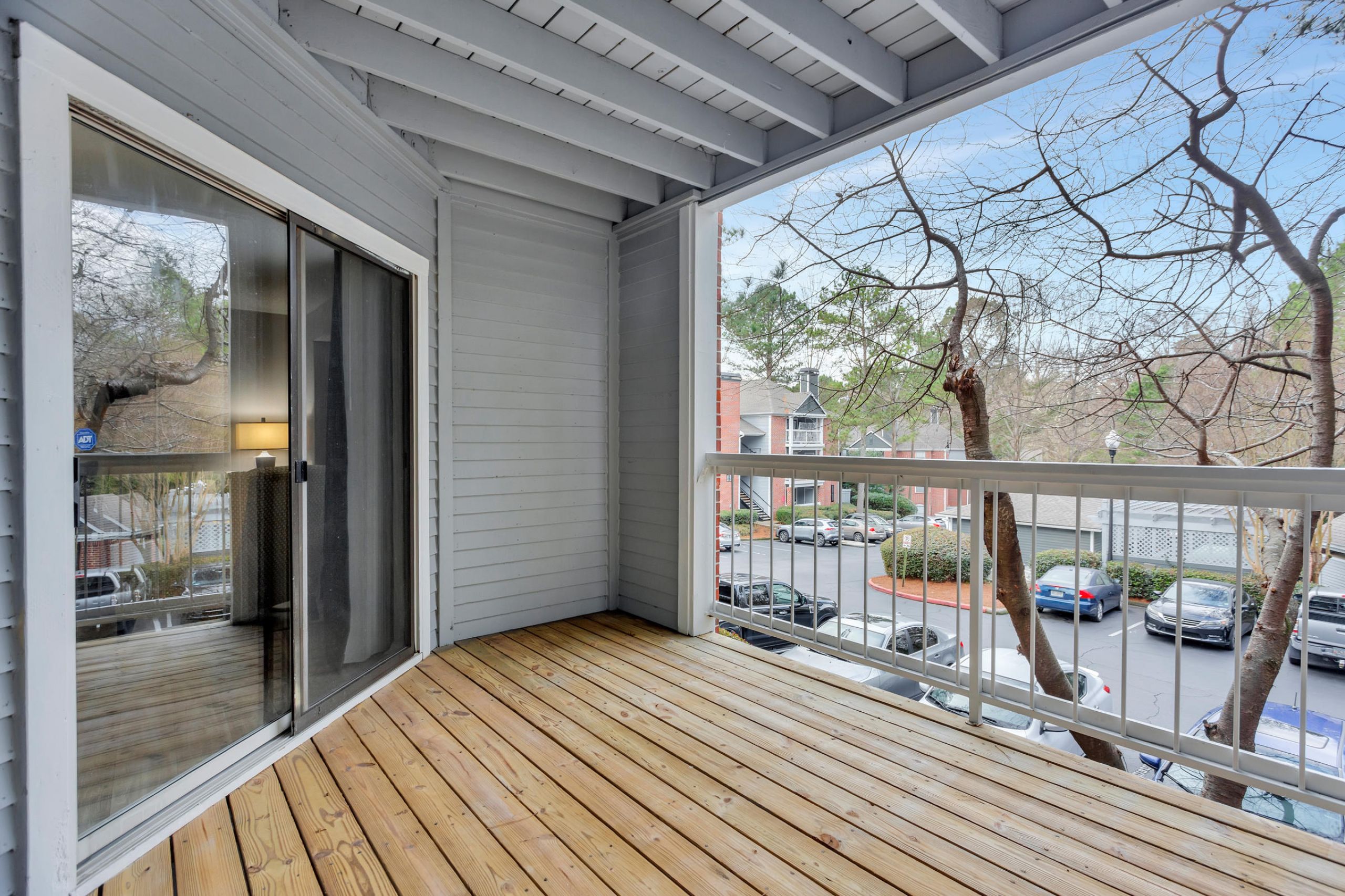 Second-floor balcony with wooden floor, glass sliding door, and view of parking lot and trees in an apartment complex.