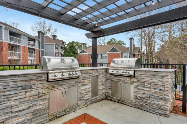 Outdoor kitchen with two stainless steel grills under a black pergola, surrounded by apartment buildings.