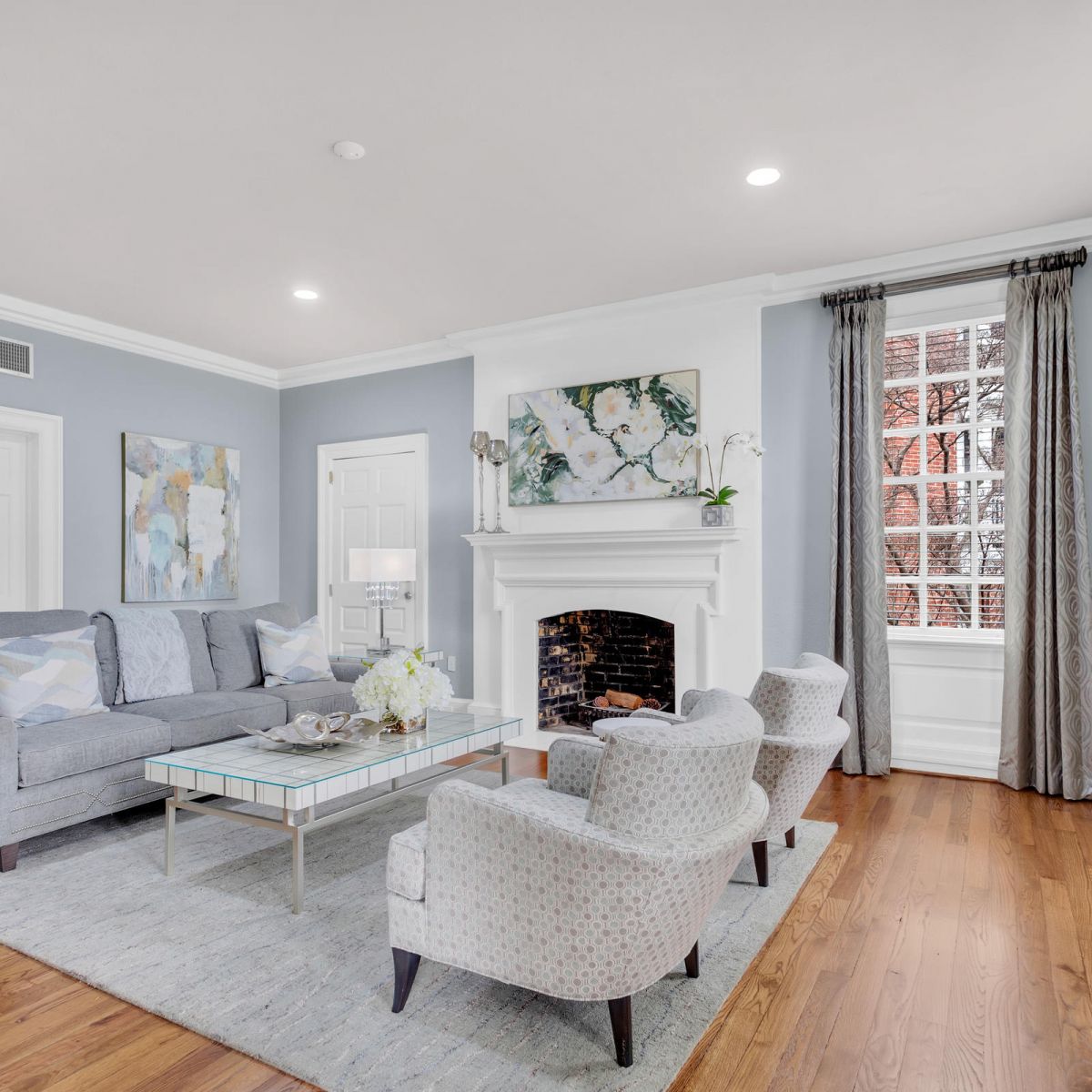 Bright living room with gray furniture, a fireplace, art on the walls, large windows, and hardwood floors.