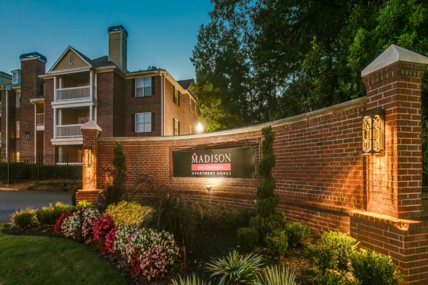 Brick apartment complex at dusk with a lit "Madison Brookhaven Apartment Homes" sign and colorful flowers in front.