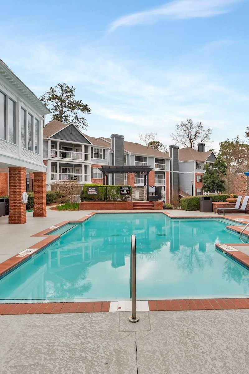 Outdoor swimming pool with steps, surrounded by lounge chairs and modern apartment buildings in the background.