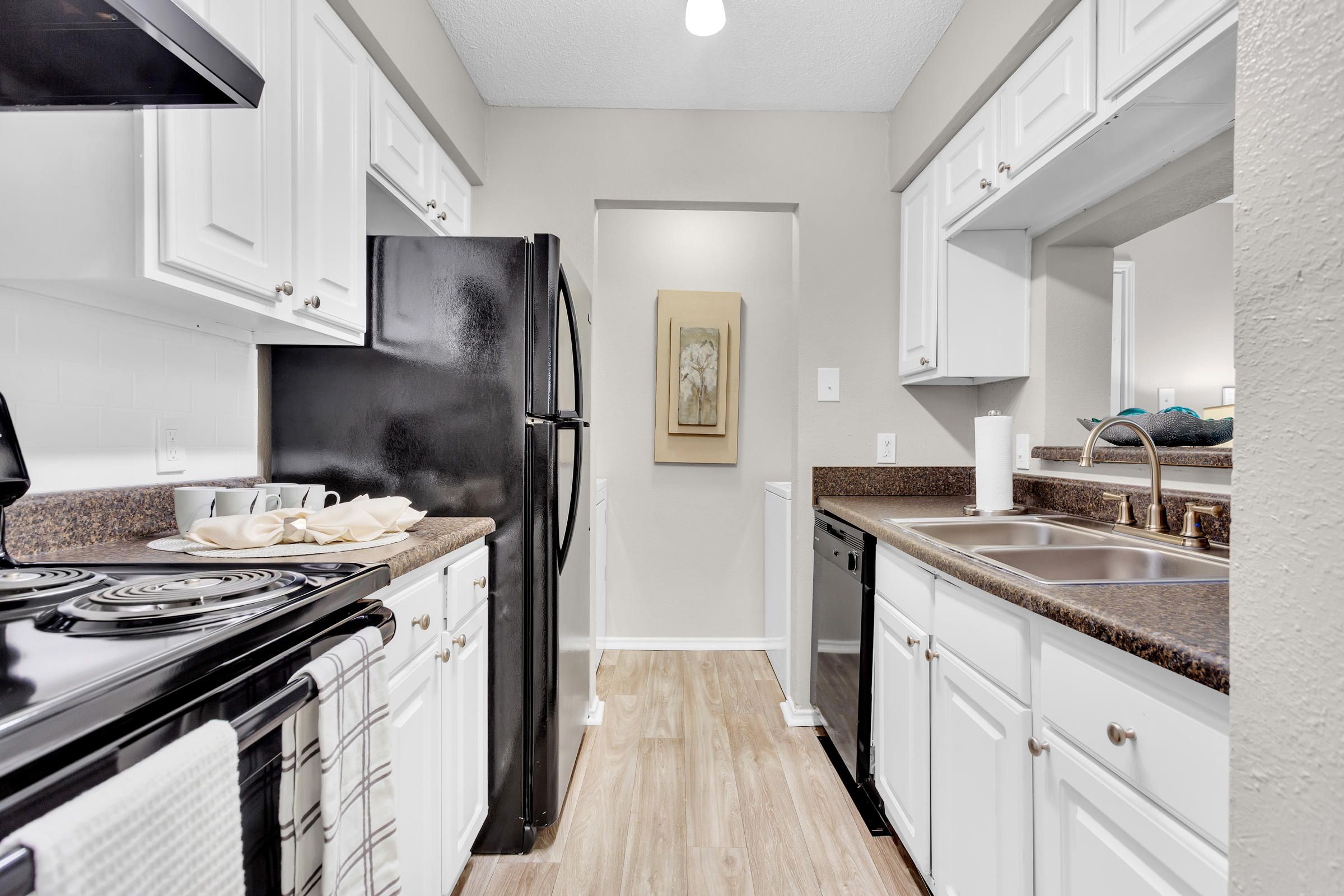 Modern galley kitchen with white cabinets, black appliances, and brown countertops, viewed from the entrance.