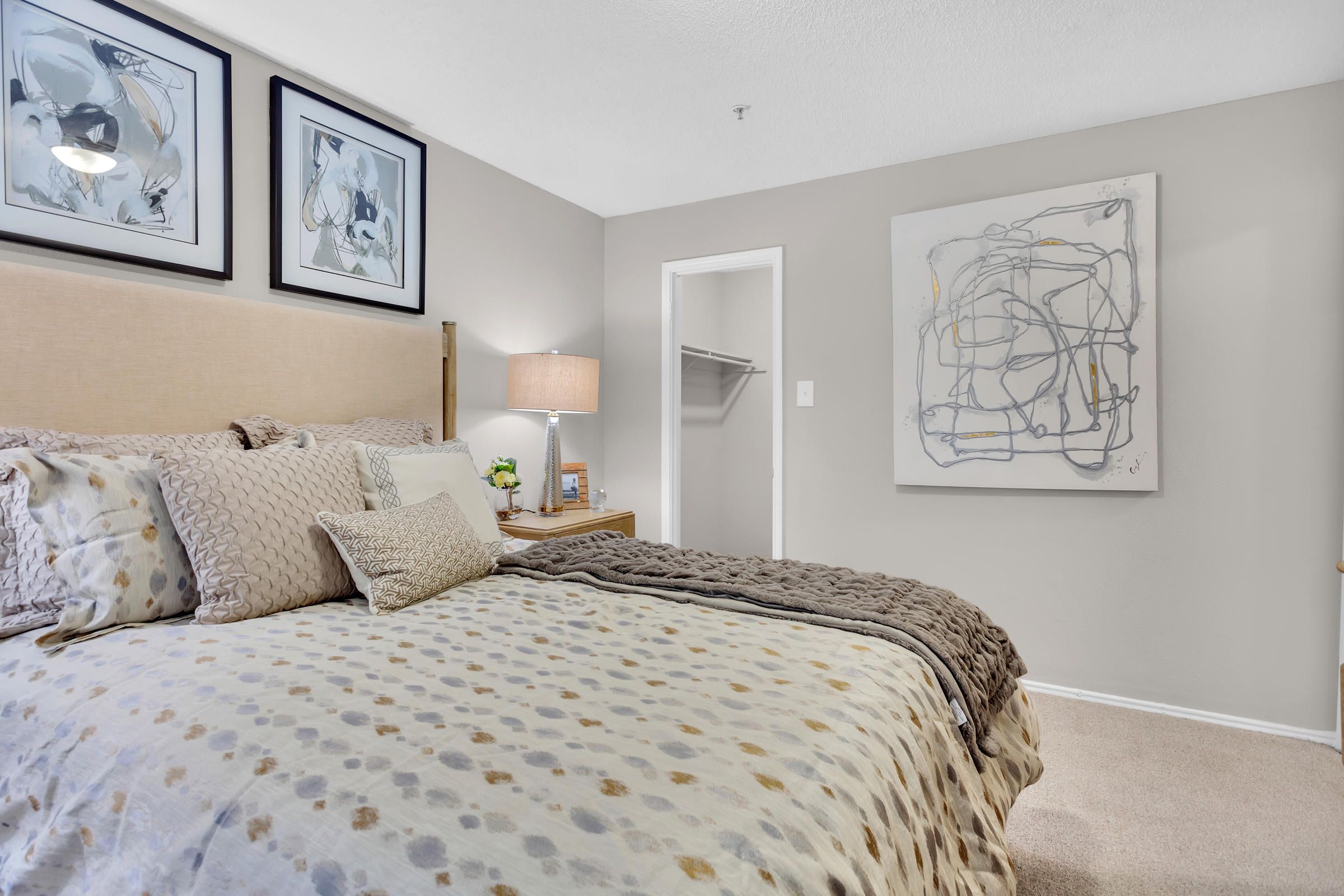 Cozy bedroom with neutral bedding, abstract wall art, a lamp on a wooden nightstand, and open closet door.