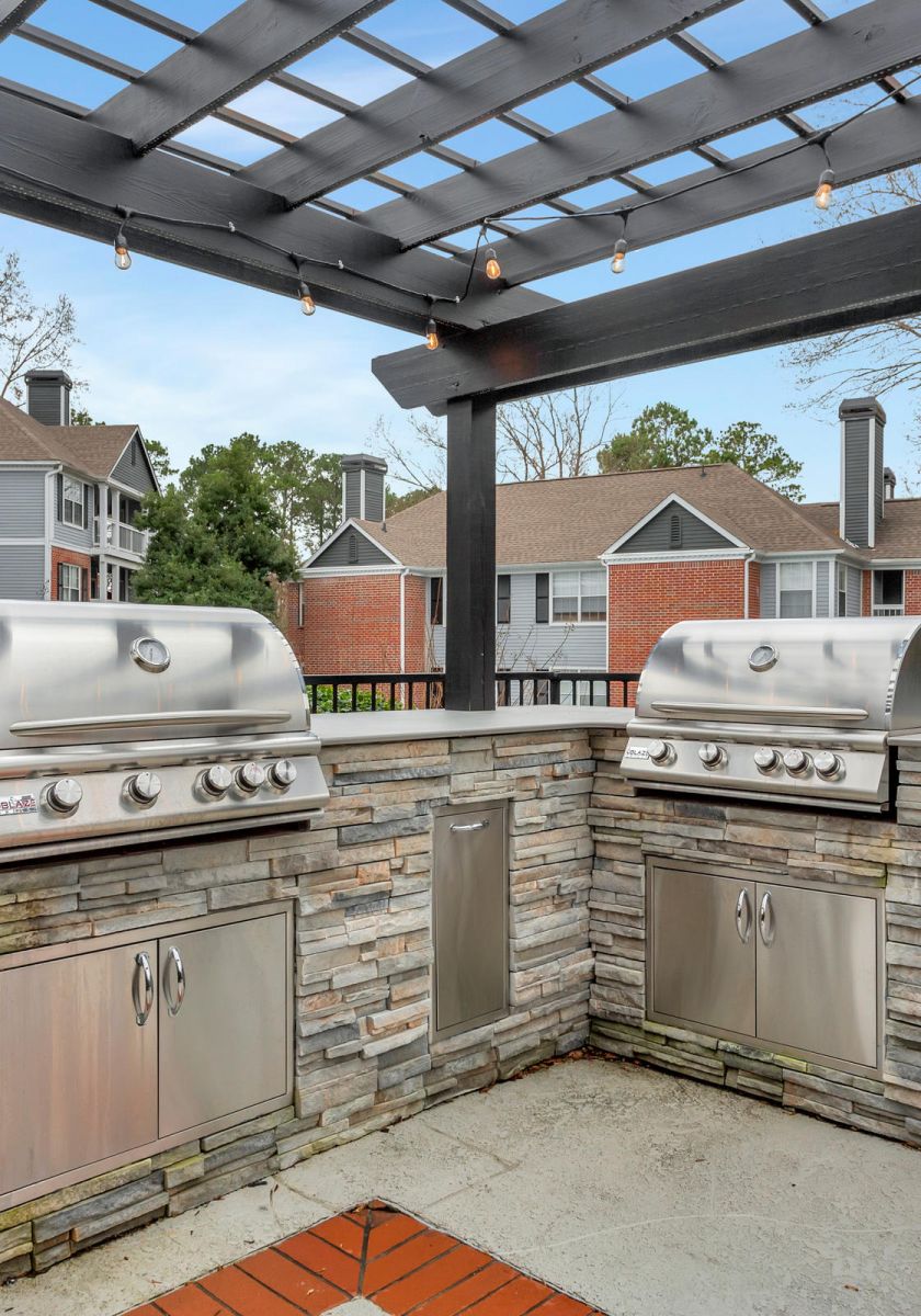 Outdoor kitchen with two stainless steel grills under a black pergola, surrounded by apartment buildings.