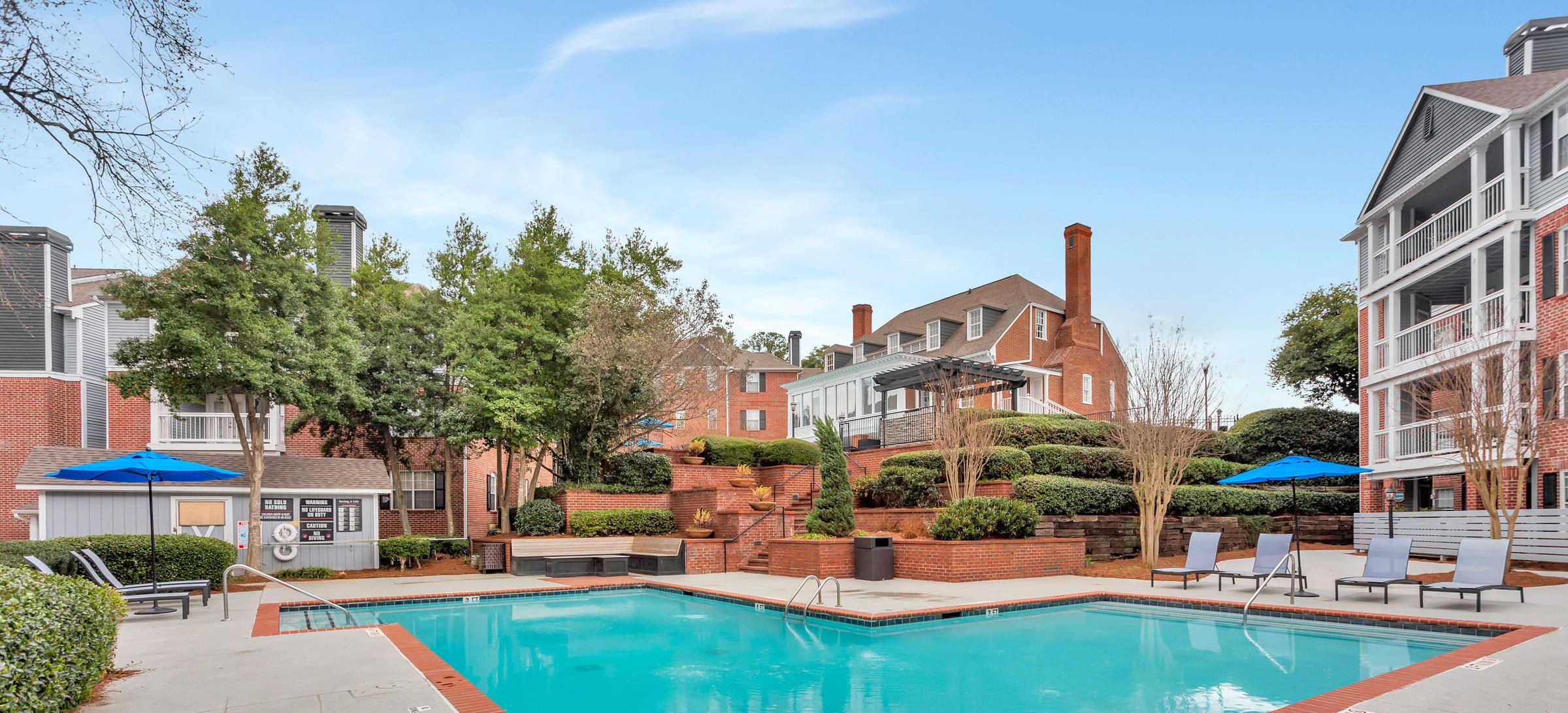 Outdoor swimming pool with lounge chairs, umbrellas, and red-brick apartment buildings in the background.