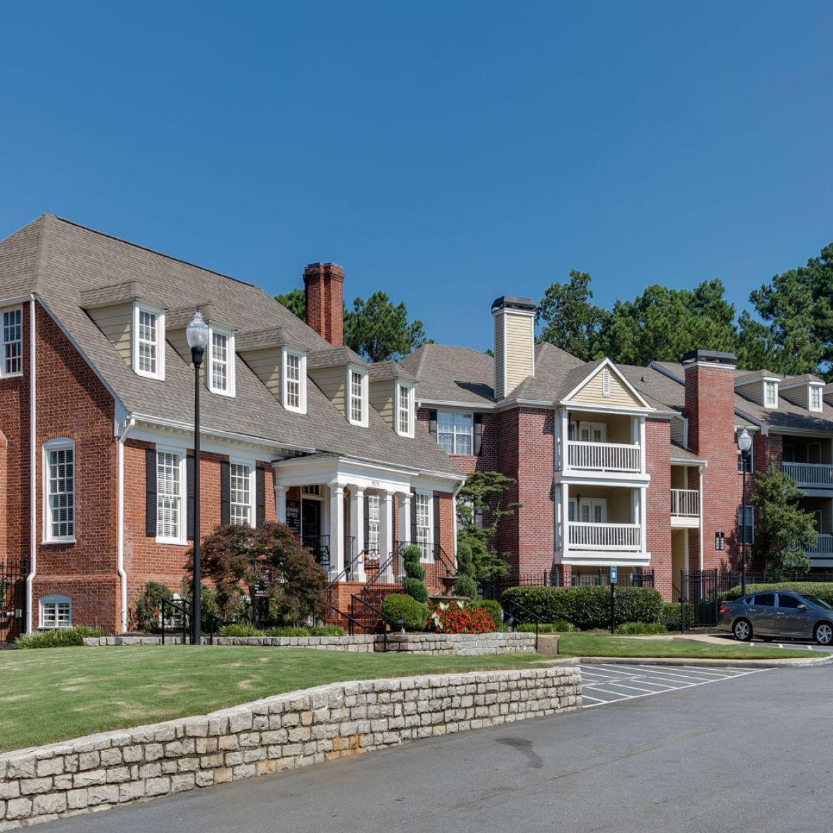 Red-brick apartment buildings with white trim and balconies, trees behind, and cars parked in front.