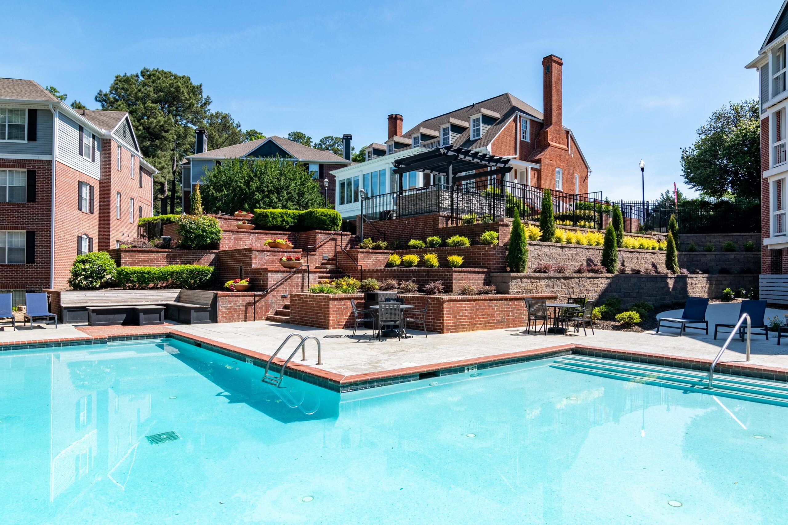 Outdoor swimming pool with clear blue water, surrounded by patio seating and multi-story brick buildings.