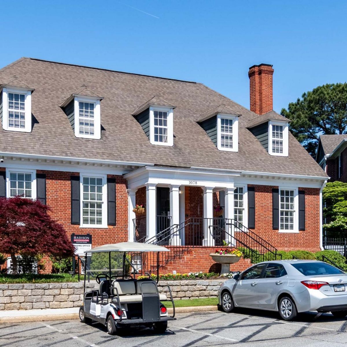 A brick house with a steep roof, dormer windows, and white columns, with cars parked in front.
