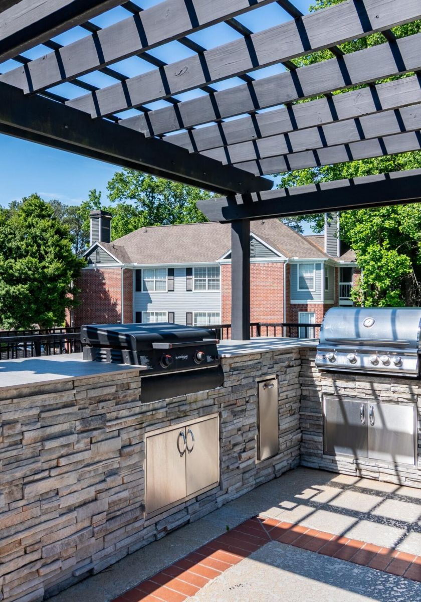 Outdoor kitchen with stone counters, two grills, and a pergola, surrounded by apartment buildings and trees.